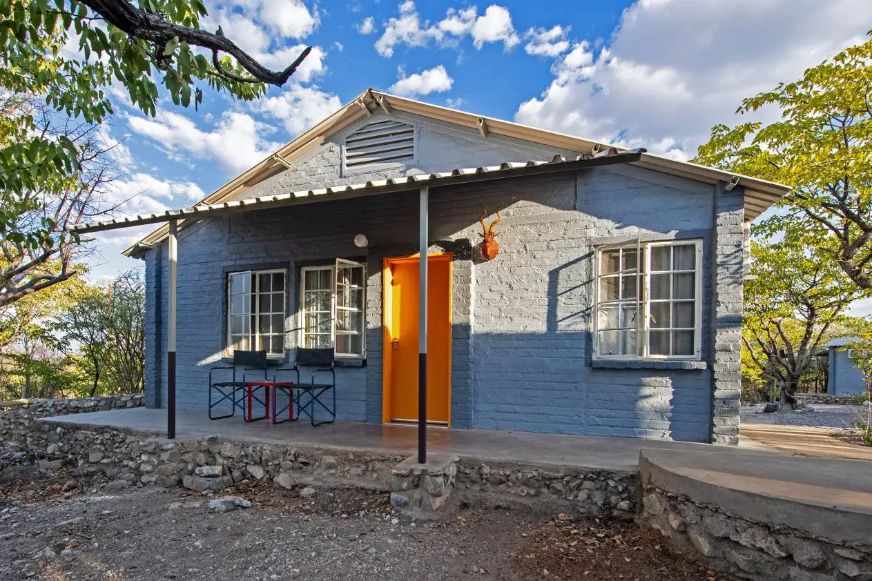 Blue-painted brick chalet with a bright orange door and shaded veranda at Etosha Safari Camp