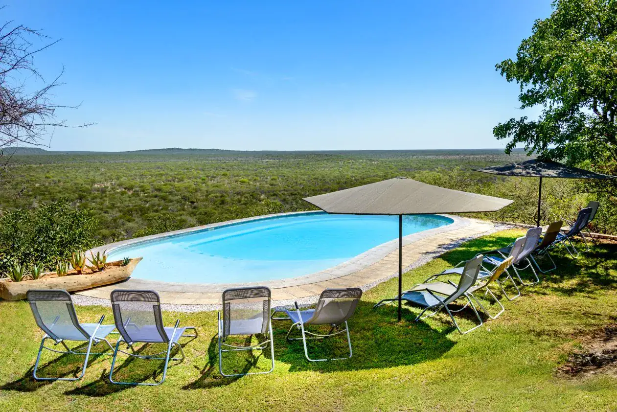 Swimming pool with umbrellas and loungers on the hilltop lawn overlooking mopane woodland at Etosha Safari Lodge