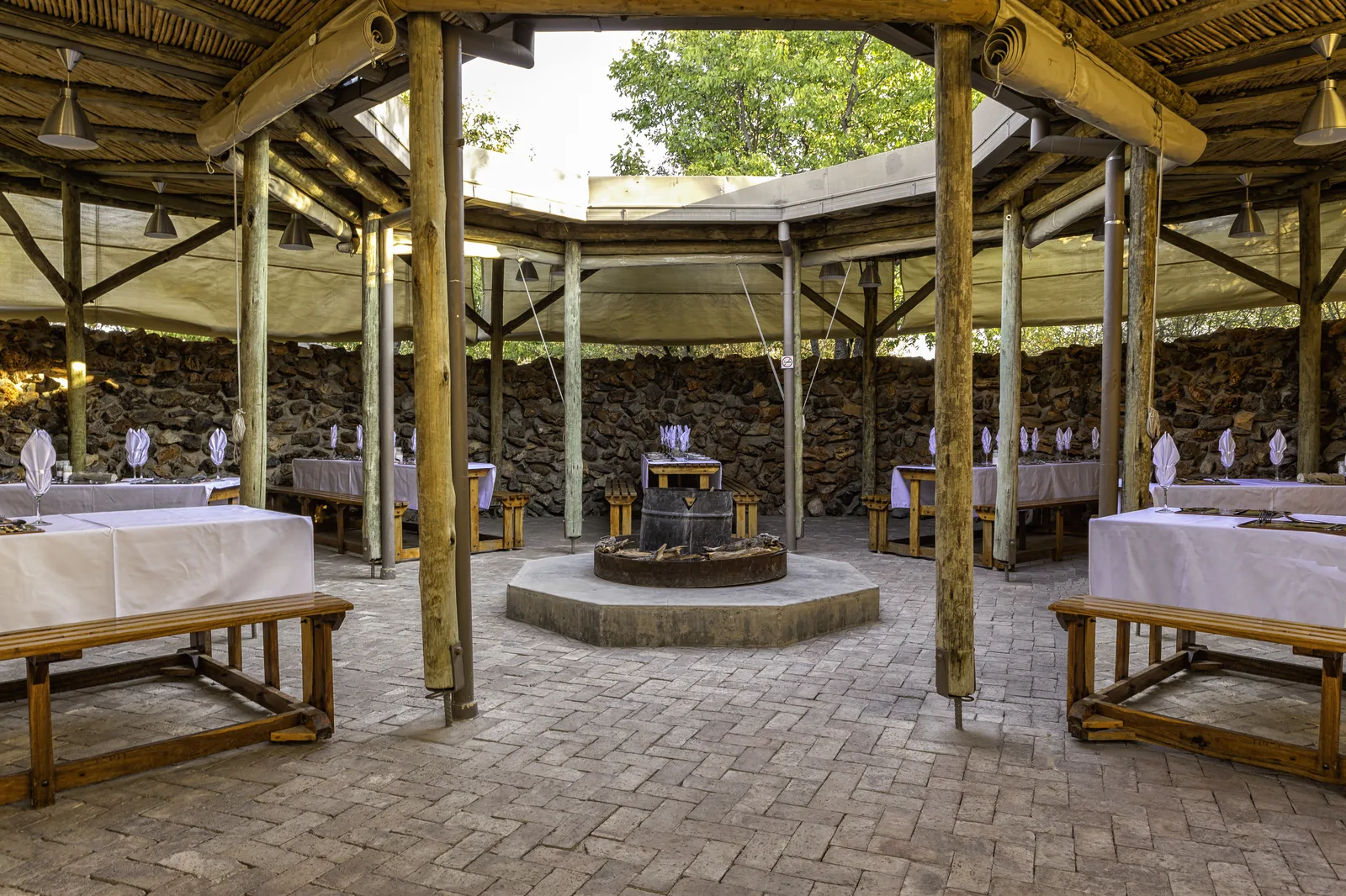 Covered open-air boma dining area with a central firepit and set tables at Etosha Village