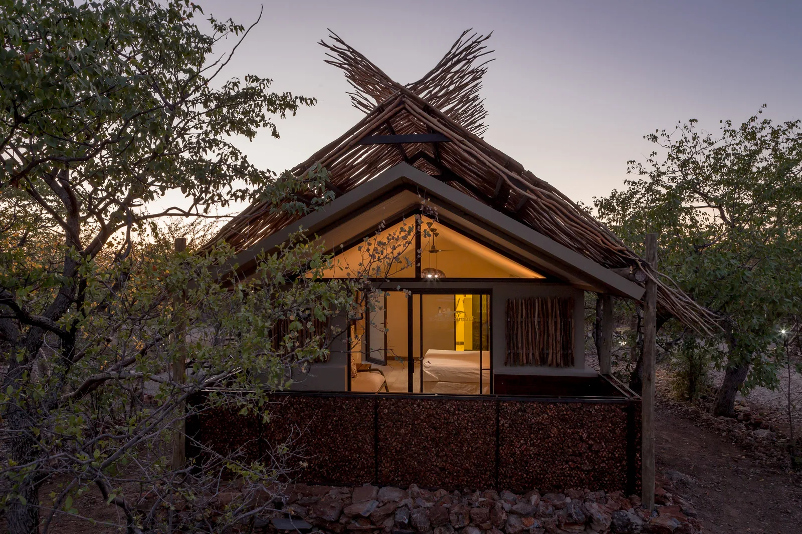 A glowing thatched-roof tented unit at dusk among mopane trees at Etosha Village, 2 km from Etosha National Park