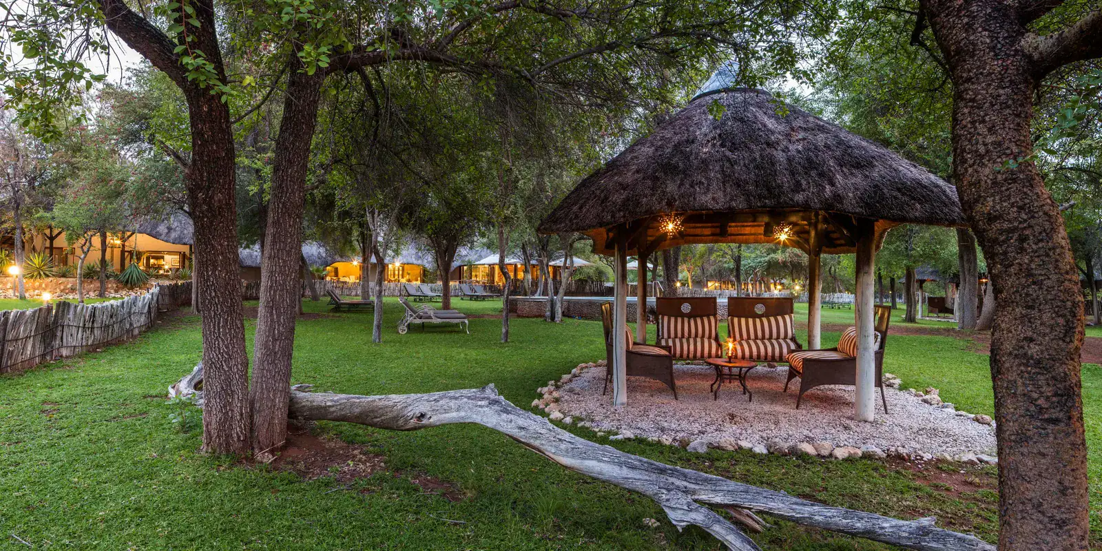 Thatched garden gazebo and lawns at dusk with the pool and lamp-lit chalets of Mushara Lodge behind